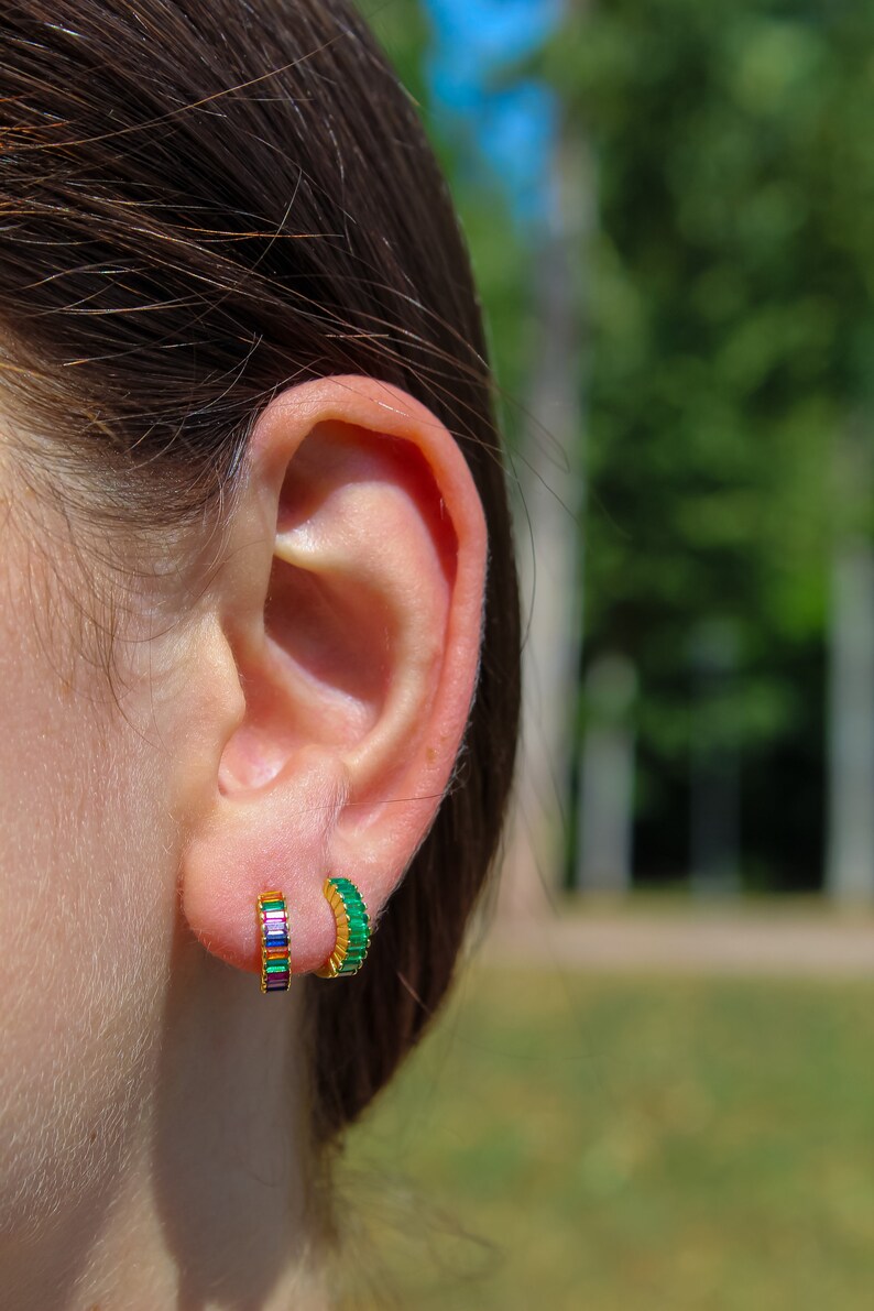 May include: A close-up of a person's ear with two gold hoop earrings. The earring on the left is set with multi-colored stones, while the earring on the right is set with green stones.