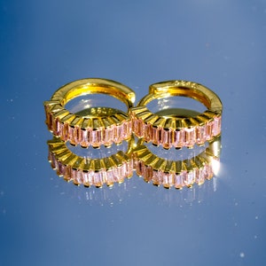 May include: A pair of gold hoop earrings with pink baguette-shaped stones. The earrings are reflected in a blue surface.