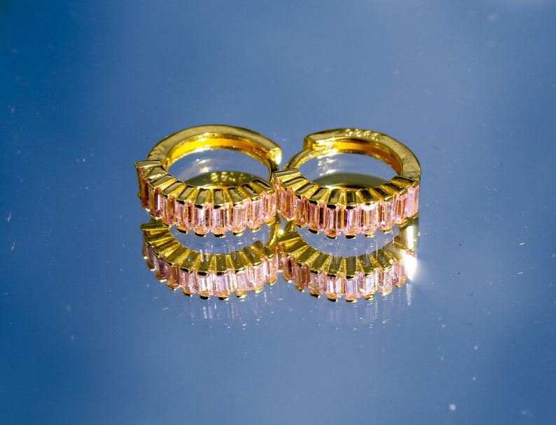 May include: A pair of gold hoop earrings with pink baguette-shaped stones. The earrings are reflected in a blue surface.