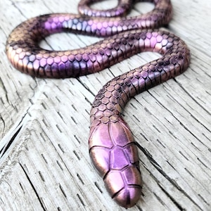 May include: A decorative snake figurine with a metallic finish. The snake is primarily purple with bronze and pink highlights, and features a scale pattern. The figurine is posed on a wooden surface.