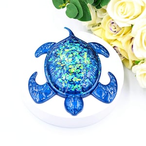Peut inclure: Une tortue en résine bleue avec une carapace scintillante verte et bleue. La tortue est posée sur une base ronde blanche.