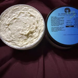 Puede incluir: Un tarro abierto de Edge Growth Butter de 12 Tribes on Earth, con una textura cremosa blanquecina. La tapa, con una etiqueta azul y negra, enumera los ingredientes e instrucciones. El producto est&aacute; dise&ntilde;ado para hacer crecer el cabello de forma natural.