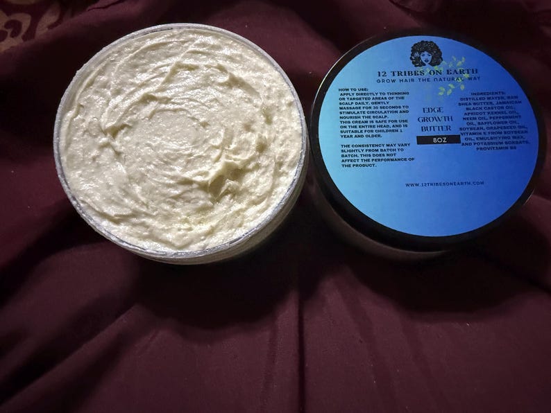 Puede incluir: Un tarro abierto de Edge Growth Butter de 12 Tribes on Earth, con una textura cremosa blanquecina. La tapa, con una etiqueta azul y negra, enumera los ingredientes e instrucciones. El producto est&aacute; dise&ntilde;ado para hacer crecer el cabello de forma natural.