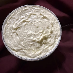 Puede incluir: Vista cenital de un recipiente redondo lleno de una crema espesa de color blanquecino. La crema tiene un aspecto texturizado y arremolinado. El recipiente est&aacute; sobre un fondo de tela burdeos oscuro.