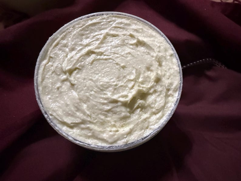 Puede incluir: Vista cenital de un recipiente redondo lleno de una crema espesa de color blanquecino. La crema tiene un aspecto texturizado y arremolinado. El recipiente est&aacute; sobre un fondo de tela burdeos oscuro.