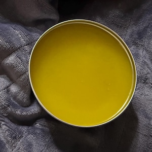 May include: A round metal tin filled with a smooth, solid, olive-green balm. The container has a simple, circular silver-colored rim that frames the contents, which appear to have a consistent, waxy texture throughout the entire surface area.