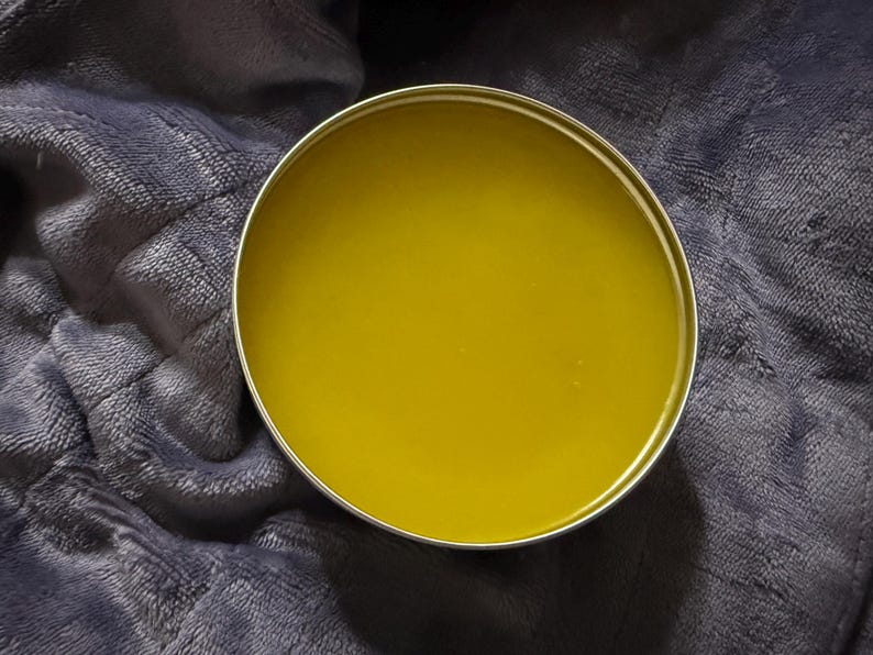 May include: A round metal tin filled with a smooth, solid, olive-green balm. The container has a simple, circular silver-colored rim that frames the contents, which appear to have a consistent, waxy texture throughout the entire surface area.