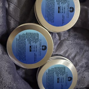 May include: Three round gold-colored metal tins of 12 Tribes on Earth Creamy Emu Oil Pomade. Each lid features a blue circular label with black text listing ingredients including emu oil, vanilla essence, peppermint oil, and beeswax, plus 8oz weight.