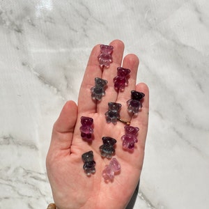 May include: A hand holding a collection of small, clear glass teddy bear figurines in various shades of purple, pink, and gray.