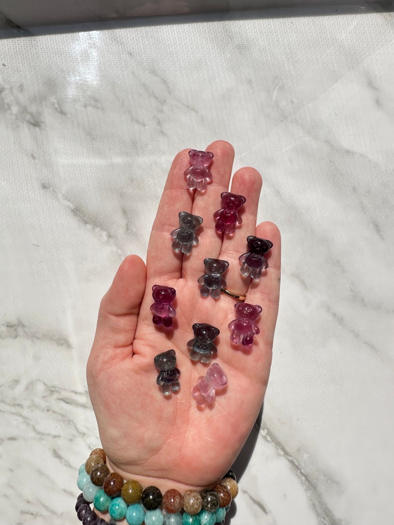 May include: A hand holding a collection of small, clear glass teddy bear figurines in various shades of purple, pink, and gray.