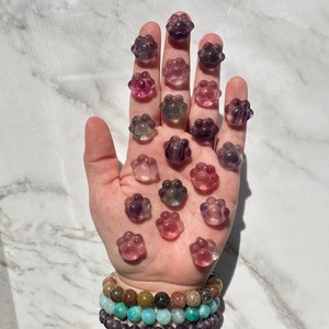 May include: A hand holding a collection of 20 small, purple, pink, and blue paw print shaped crystal charms.