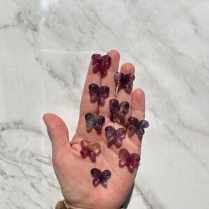 May include: A collection of purple, pink, and blue butterfly-shaped fluorite crystals. The crystals are arranged on a hand, with the hand wearing a multi-colored beaded bracelet.