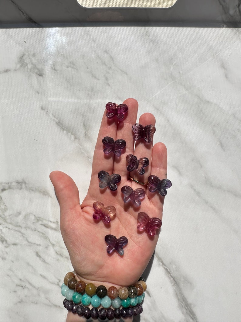 May include: A collection of purple, pink, and blue butterfly-shaped fluorite crystals. The crystals are arranged on a hand, with the hand wearing a multi-colored beaded bracelet.