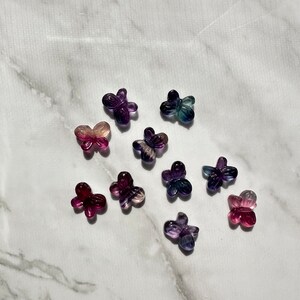 May include: A collection of ten small butterfly-shaped charms made of iridescent fluorite in shades of purple, pink, and blue. The charms are arranged on a white marble surface.