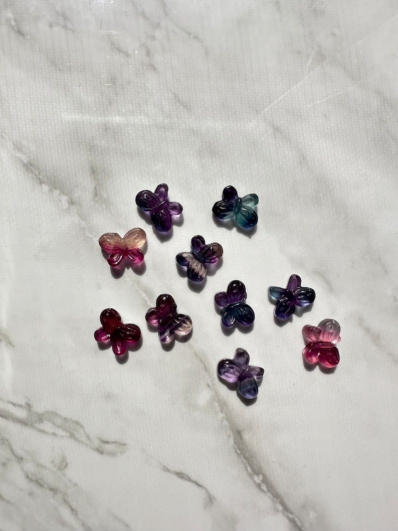 May include: A collection of ten small butterfly-shaped charms made of iridescent fluorite in shades of purple, pink, and blue. The charms are arranged on a white marble surface.