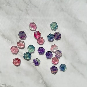 May include: Assortment of colorful paw print shaped resin charms. The charms are translucent and have a variety of colors including pink, purple, blue, and green.