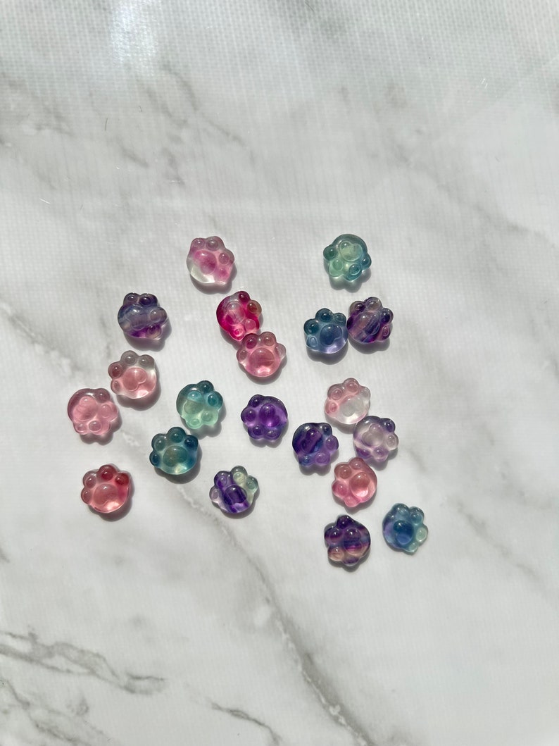 May include: Assortment of colorful paw print shaped resin charms. The charms are translucent and have a variety of colors including pink, purple, blue, and green.