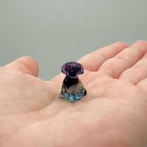 May include: A small, glass figurine of a dog sitting. The figurine is made of clear glass with a purple and blue iridescent finish.