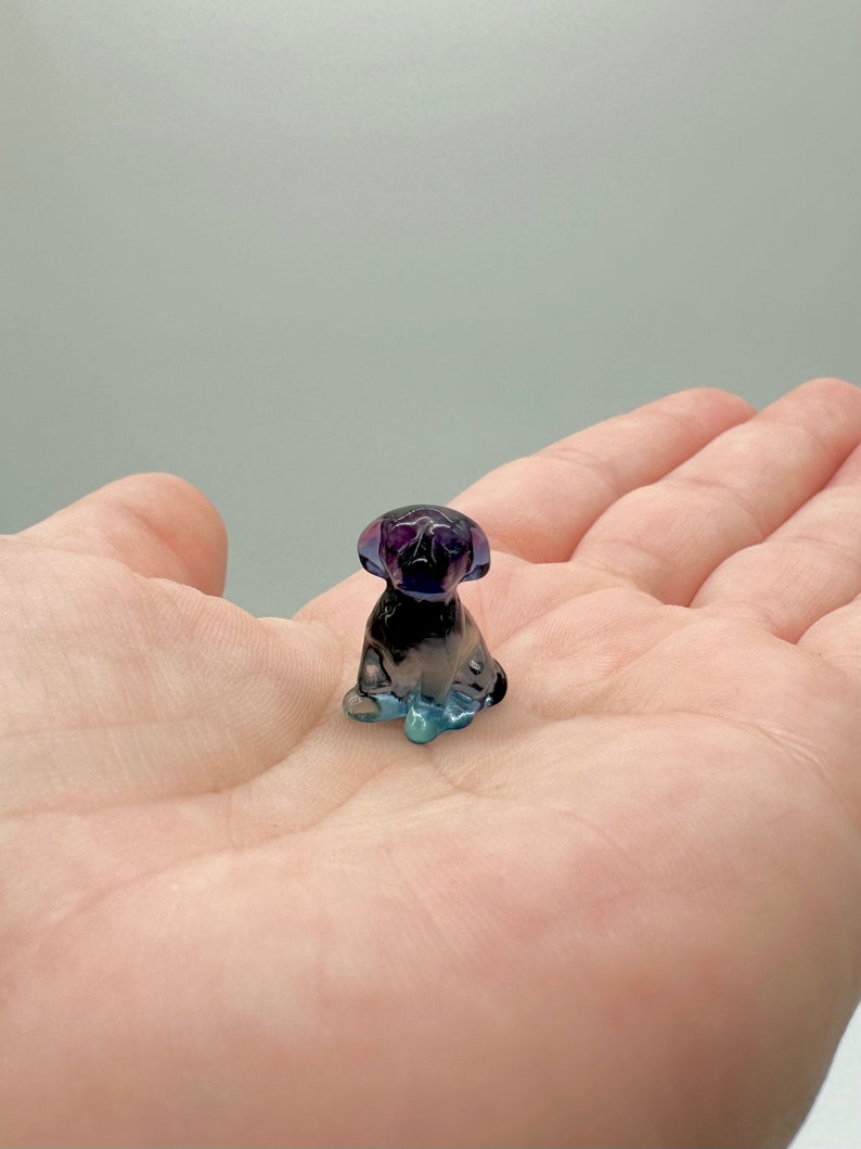 May include: A small, glass figurine of a dog sitting. The figurine is made of clear glass with a purple and blue iridescent finish.