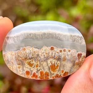 Könnte beinhalten: Ein ovales Schmuckstück mit Landschaftsdesign. Der obere Teil ist hellblau, geht in ein weißes und graues Band über und dann in eine Landschaft aus braunen und orangefarbenen Formationen. Der Stein wird zwischen zwei Fingern gehalten.