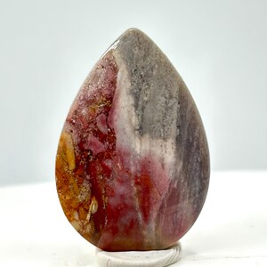 May include: A polished, teardrop-shaped stone displaying a blend of red, orange, grey, and white hues. It is mounted on a small, white, oval base. The stone's surface has a smooth, glossy finish, showcasing natural patterns.