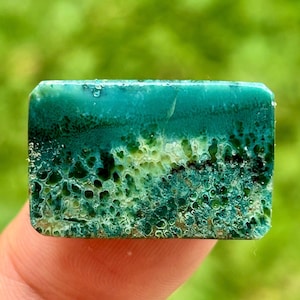 Könnte beinhalten: Ein rechteckiger, polierter Edelstein mit geschichtetem Aussehen. Die obere Schicht ist ein tiefes Türkis, das in eine strukturierte, poröse Oberfläche mit Grün- und Gelbtönen übergeht. Der Stein wird von einem Finger vor einem verschwommenen grünen Hintergrund gehalten.