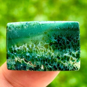 Könnte beinhalten: Ein rechteckiger Edelstein in leuchtendem Grün, mit einer Mischung aus hellen und dunklen Farbtönen. Der Stein hat ein einzigartiges Muster mit einer strukturierten Oberfläche, die grüne und weiße Farbtöne kombiniert. Der Edelstein wird von einem Finger gehalten.
