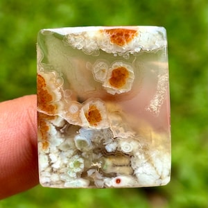 Könnte beinhalten: Ein rechteckiger Edelstein mit transluzenter Basis, der komplizierte Muster aus Weiß, Orange und Braun aufweist. Die Oberfläche des Steins hat ein wolkiges, geschichtetes Aussehen mit poliertem Finish. Der Edelstein wird vor einem verschwommenen grünen Hintergrund gehalten.