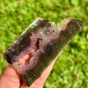 May include: A rectangular, natural agate stone with a mossy green exterior and a translucent, light purple interior. The stone has a jagged edge and a central cavity, showcasing its unique geological formation.