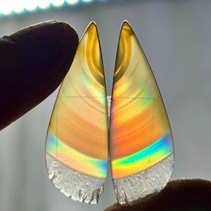 Puede incluir: Dos gemas translúcidas en forma de lágrima con iridiscencia de arcoíris. Las piedras tienen un degradado de colores, incluyendo amarillo, naranja y azul. Los bordes inferiores son ásperos y cristalinos. Las gemas se sostienen entre los dedos.