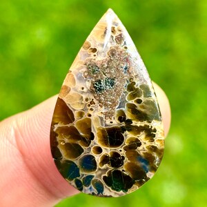 Könnte beinhalten: Ein tropfenförmiger Edelstein mit einem einzigartigen Muster aus Braun-, Beige- und Dunkelgrüntönen. Die polierte Oberfläche zeigt ein komplexes, organisches Design mit einer Mischung aus Texturen und Farben, ideal für die Schmuckherstellung oder das Sammeln.