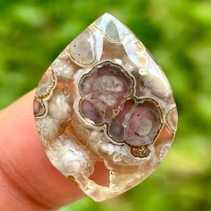 Könnte beinhalten: Ein polierter, tropfenförmiger Edelstein mit einem einzigartigen Muster. Der Stein weist eine Mischung aus Farben auf, darunter Rosa-, Braun- und Weißtöne, mit einer durchscheinenden Qualität. Das komplizierte Design ähnelt einer Reihe von geschichteten Formationen.