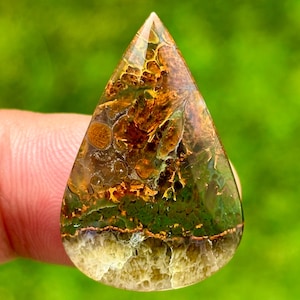 Könnte beinhalten: Ein tropfenförmiger Edelstein mit polierter Oberfläche. Der Stein weist eine Mischung aus Farben auf, darunter Braun, Grün und Gelb, mit komplizierten Mustern, die einer Landschaft ähneln. Der Edelstein wird vor einem verschwommenen grünen Hintergrund gehalten.