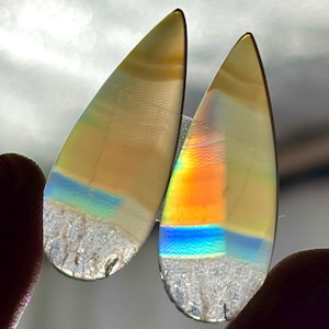Puede incluir: Dos piedras preciosas iridiscentes en forma de lágrima. Las piedras tienen un tono amarillo claro translúcido con bandas de colores naranja, azul y arcoíris. Los bordes inferiores tienen una apariencia texturizada y cristalina.