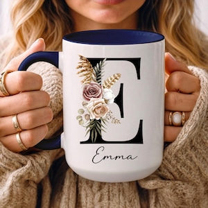 May include: White ceramic mug with a navy blue interior and handle. The mug features a large black letter "E" adorned with floral and feather accents. The name "Emma" is written in script below the letter.
