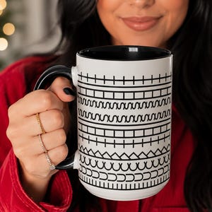 May include: A white ceramic mug with a black interior and a black handle, featuring a repeating black geometric pattern of horizontal lines, squares, wavy lines, and triangles wrapped around the exterior surface of the cylindrical vessel.
