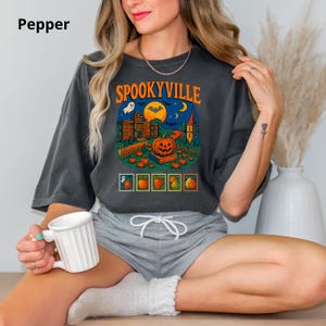 May include: A charcoal gray t-shirt featuring the text SPOOKYVILLE in orange letters above a graphic of a haunted house, a large pumpkin, bats, and a moon. Below the main scene are five small rectangular frames containing fruit illustrations.
