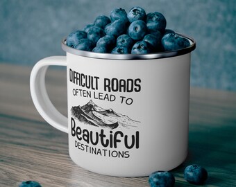 May include: A white enamel mug filled with fresh blueberries. The mug features a black mountain graphic and the text: DIFFICULT ROADS OFTEN LEAD TO BEAUTIFUL DESTINATIONS. The design is printed in a bold, dark font on the front of the smooth white surface.