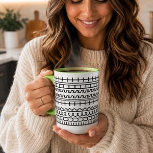 May include: A white ceramic mug featuring a repeating black geometric pattern of horizontal lines, squares, and zigzags. The mug has a bright lime green handle and a matching lime green interior rim, containing a dark steaming hot beverage.