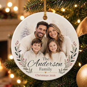 May include: A round white ceramic Christmas ornament featuring a printed photograph of a family of four wearing cream sweaters. The ornament is decorated with green pine branches and red berries, with the text The Anderson Family Christmas 2026.