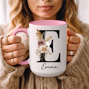Puede incluir: Taza de cerámica blanca con interior y asa rosas. La taza presenta una gran letra negra "E" adornada con elementos florales y botánicos. El nombre "Emma" está escrito en cursiva debajo.