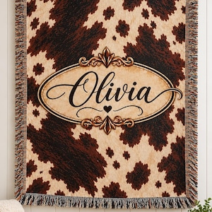 May include: A woven throw blanket featuring a brown and cream cowhide print pattern. In the center, an oval frame with decorative flourishes surrounds the name Olivia written in black script, with a small heart icon below the name.