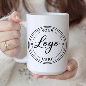 May include: A white ceramic mug featuring a circular black graphic design on the front. The design consists of a double-lined circle with diamond accents and the text YOUR Logo HERE printed in a combination of serif and script fonts.