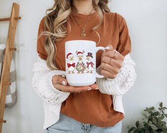 Op de afbeelding: Een witte keramische mok met een afbeelding van drie ganzen die zijn aangekleed voor de feestdagen. Eén gans draagt een kerstmuts en rode vlinderdas, de middelste gans heeft rendiergeweien en kerstverlichting, en de derde een hoge hoed.