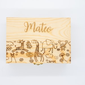 Puede incluir: Una caja de madera de color claro con el nombre "Mateo" grabado en la parte superior. La caja presenta un borde decorativo con ilustraciones de animales grabadas, incluyendo una cebra, una jirafa, un elefante y un koala.