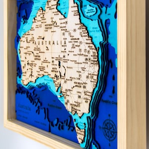 May include: A framed, three-dimensional wooden map of Australia. The map features a light wood landmass and blue ocean layers. The map is encased in a natural wood frame, with measurements in centimetres.