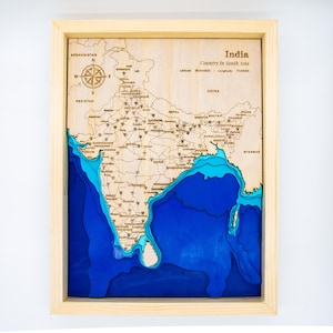 May include: Wooden framed map of India, laser-cut with detailed topography. The map features a compass rose and text indicating the country's name and location in South Asia. The ocean is depicted in shades of blue.