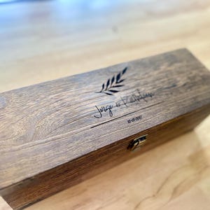 May include: A wooden box with a hinged lid and metal clasp. The box is engraved with the names "Jorge & Kathlyn", a sprig design, and the date "10-01-2017". The wood has a natural grain and a warm brown tone.