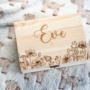 Peut inclure: Une boîte en bois de couleur claire avec le nom "Eve" gravé sur le couvercle. La boîte présente un motif floral le long du bord inférieur. La boîte repose sur une couverture douce et à motifs.