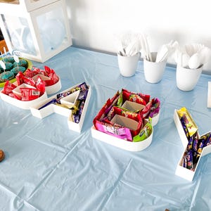 May include: A party table with the word "A-Y-A-Y" spelled out in white containers filled with sweets. Cupcakes with blue icing and a wooden board with green treats are also on the table. White cups with cutlery are also present.